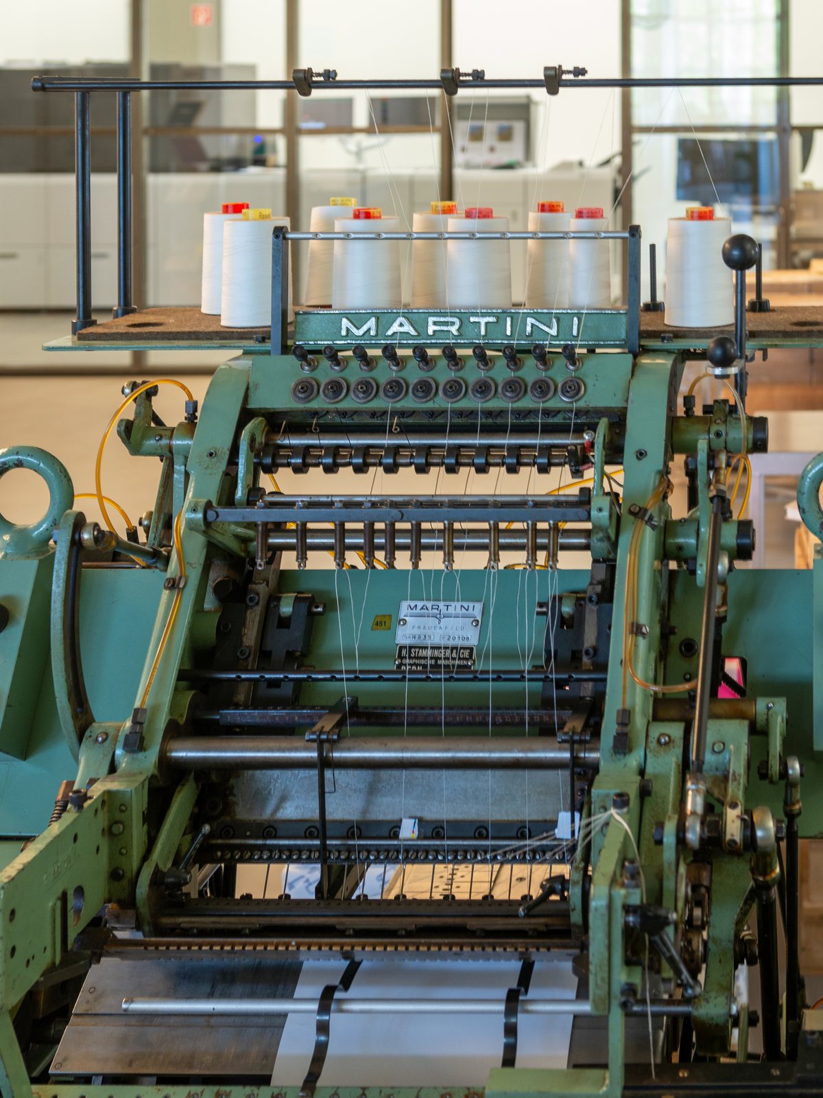 Fadenheftmaschine Martini mit aufgesetzten Garnrollen in der Buchbinderei an der Uferstrasse 90, dahinter durch die Glaswand der Digitaldruck